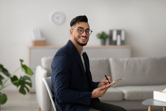 Male Arab psychotherapist with a clipboard smiles at the camera in a modern office. He appears approachable and professional while providing psychological services to clients.