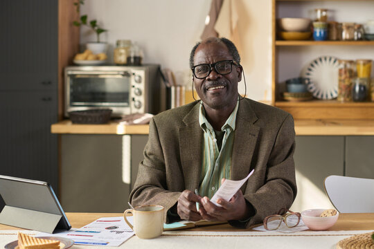 Portrait of senior Black man wearing glasses smiling while sitting at kitchen table holding documents and listening to earphones, digital tablet and paperwork on table