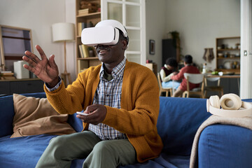 Mature Black man sitting on sofa using virtual reality headset and handheld controller, gesturing with hand while interacting with digital environment, family in background