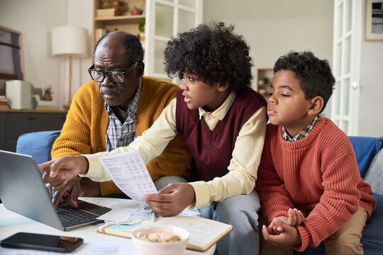 Black senior man sitting with two Black boys, one teenager and one child, working together on laptop and reviewing financial documents, teenager explaining information to others
