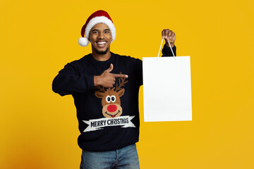 A cheerful man in a Santa hat smiles broadly while holding an empty gift bag. He points to the bag, ready for Christmas celebrations. The background is a vibrant yellow, adding to the festive mood.