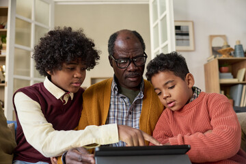 Senior Black man sitting between two Black boys using digital tablet together, boys focusing on screen while man supervising, multigenerational family engaging with technology