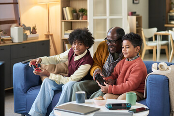 Black senior man sitting on sofa playing video game with two Black boys, both children, all smiling and looking at television screen, holding game controllers in living room setting