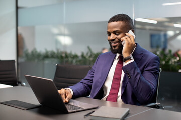 Successful african american young businessman in expensive blue suit working on laptop at office, having phone conversation with his personal assistant or business partner and smiling, copy space