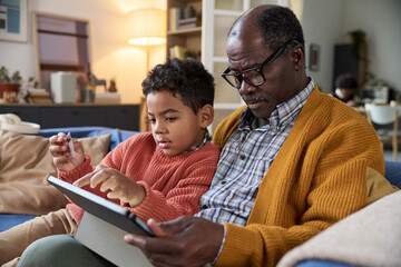 Black senior man sitting with Black child using digital tablet together on sofa, both focused on...