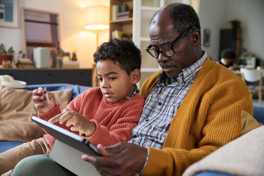 Black senior man sitting with Black child using digital tablet together on sofa, both focused on screen, child interacting with device while man supervising, cozy home background visible
