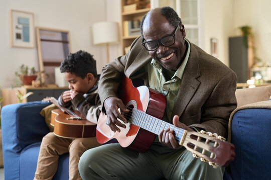 Senior Black man smiling while playing acoustic guitar, sitting on sofa with boy of Middle Eastern descent learning guitar in background, both engaging in music activity together