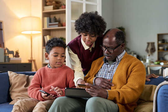 Senior man sitting on sofa using digital tablet with Black boy and Black teenager, both interacting with devices, family members engaging in technology together at home