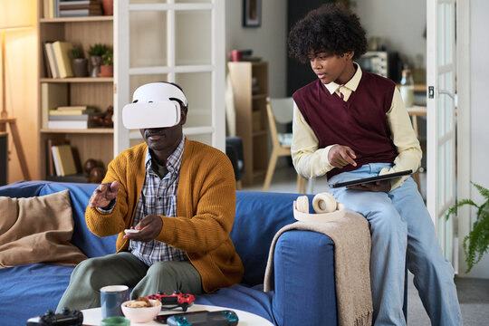 Mature Black man wearing virtual reality headset interacting with digital environment while Black teenage boy sitting nearby holding tablet observing technology use