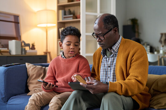 Senior Black man sitting on sofa teaching Black boy using digital tablet and holding credit card, both focused on screen, boy holding smartphone, home interior background visible