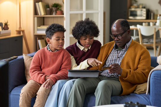 Senior Black man sitting on sofa using digital tablet with two Black boys, one child and one teenager, both attentively watching and interacting with device together