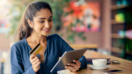 A woman joyfully engages in online shopping using a tablet while holding a credit card. She is seated at a cafe with a cup of coffee nearby, showcasing a relaxed atmosphere.