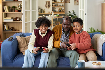 Black senior man sitting on sofa between two Black boys playing video game together, all three smiling and looking at television screen, holding game controllers in living room