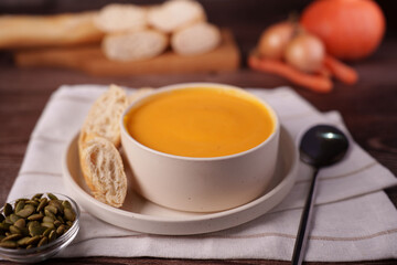 cozy autumn setup featuring a creamy pumpkin soup in a white ceramic bowl accompanied by slices of rustic bread, pumpkin seeds, and a spoon on a wooden table with soft focus background, texture
