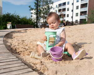 adorable toddler with playful pigtails enjoying a sunny day in a sandy urban playground, equipped with colorful sand toys, wooden paths, and modern residential buildings in the background, happy