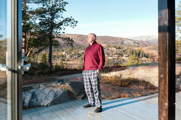 Fresh morning clarity as a senior man stands on a Nordic terrace in pajamas, looking across the...