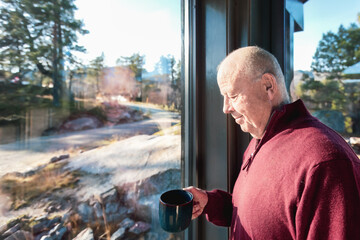 Warm morning pause as a senior man stands by a large Nordic window holding a mug, taking in the quiet landscape with slow awareness. Soft daylight, calm mood , copy space .