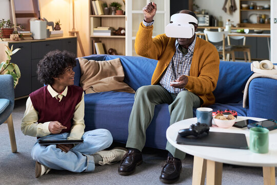 Black senior man wearing virtual reality headset sitting on sofa interacting with VR controller while Black teenage boy sitting on floor - Powered by Adobe