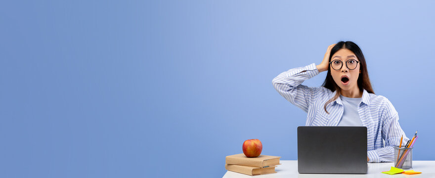 A surprised student is sitting at her desk in a blue room, looking at her laptop with wide eyes. Books and an apple are on the table beside her as she reacts with shock.