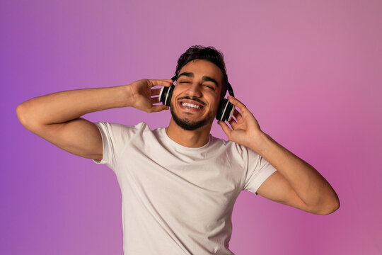 A peaceful young Arab guy listens to his favorite playlist with wireless headphones. He closes his eyes, enjoying the soothing atmosphere created by soft neon light.