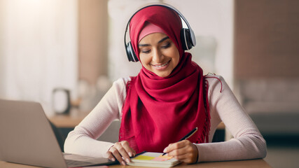 A young woman in a red hijab and headphones smiles while taking notes during a study session at her...
