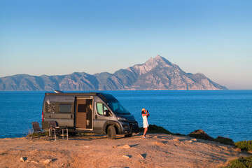 Woman enjoys the Aegean view beside a camper van with Mount Athos in the background. Ideal for: vanlife travel, outdoor brands, road-trip blogs, Greece tourism. Colours: blue, beige, silver, gold.
