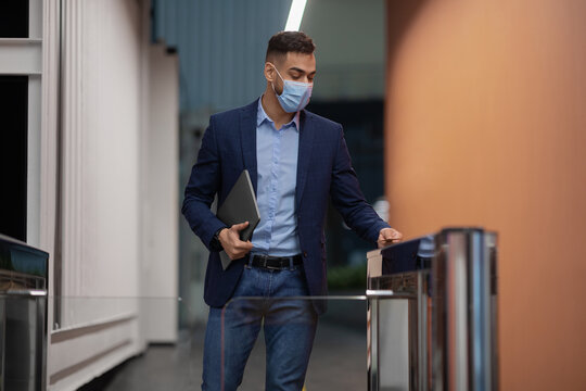 Middle eastern man wearing a protective face mask holds a laptop as he approaches the entrance gate of an office building. He uses an electronic card to gain access.