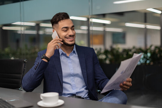 Young arab manager sits in a stylish office, reading documents with a smile. He speaks on the phone, clearly pleased with his financial report and engaged in conversation.