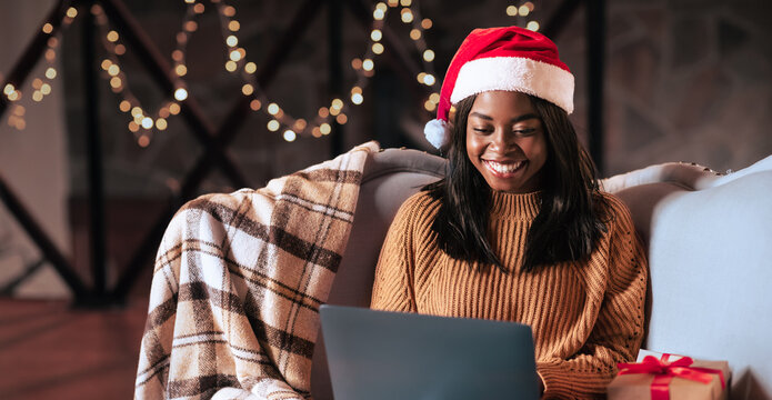 A woman sits comfortably on a cozy couch with a laptop, wearing a festive Santa hat. Holiday decorations and a gift are nearby, creating a warm Christmas atmosphere.