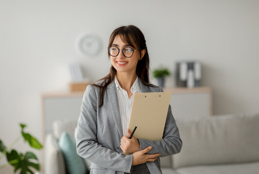 A female Arab psychotherapist stands in a modern office, smiling while holding a clipboard. She looks to the side as if engaged in thought, embodying professionalism and warmth in her environment.