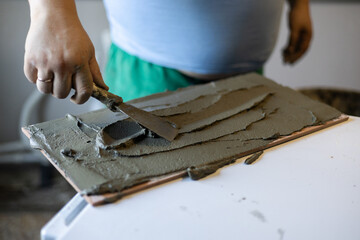 Worker's dirty hand using a metal trowel to evenly spread gray tile adhesive onto the back of a brown porcelain tile, preparing it for installation during a home renovation project.