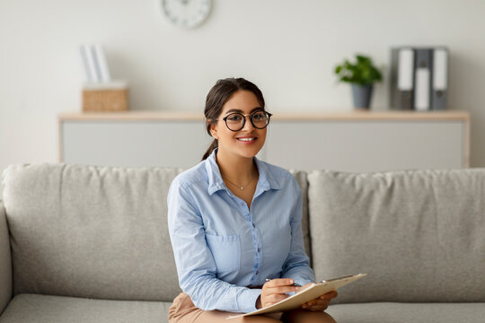 A cheerful arab female psychologist sits on a couch in her office. She is writing in her clipboard while having a session with a client, creating a friendly environment for discussion.