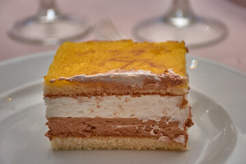 Slice of cake with layers of cream, chocolate, and yellow frosting in the foreground on a white plate in a food scene.