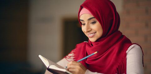 A cheerful young woman wearing a red hijab writes in her notebook with a pencil. She appears focused and content in a cozy indoor setting, immersed in her thoughts.
