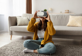 Young arab woman is seated on the floor in her living room, wearing modern VR glasses. She is fully engaged in an immersive virtual reality session at home, feeling the excitement of the experience.