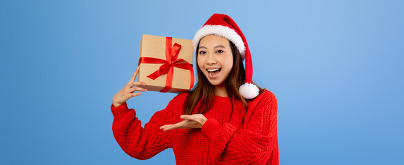 Obraz premium A young woman wears a bright red sweater and Santa hat while joyfully holding a wrapped present. She smiles widely, sharing holiday cheer in a cheerful atmosphere.
