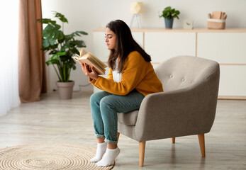 Young Arab woman enjoys a peaceful morning at home, sitting in a comfortable armchair while reading...