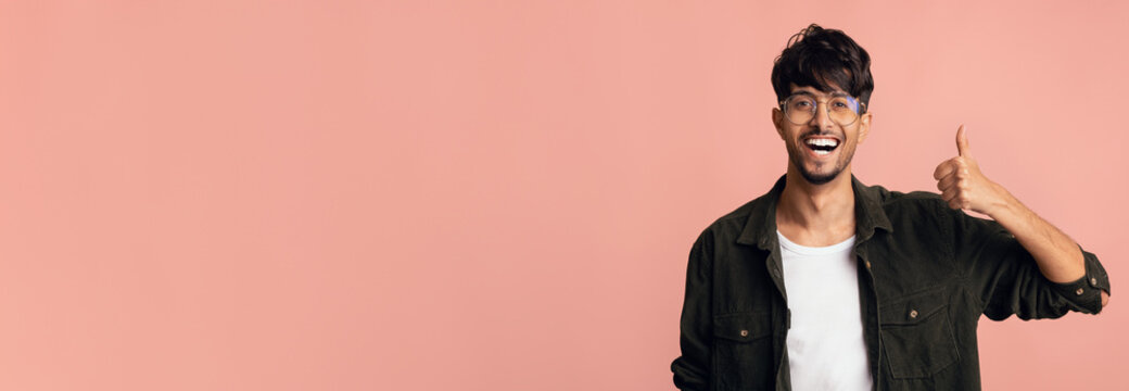 A cheerful young man stands in front of a pastel pink wall. He wears glasses and a casual outfit, smiling widely while giving a thumbs up. His energetic expression conveys positivity. - Powered by Adobe