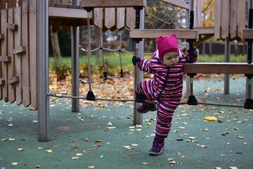 Toddler in a striped snowsuit exploring a wooden rope bridge at a playground. Autumn leaves and green flooring create a lively outdoor setting full of movement and curiosity.