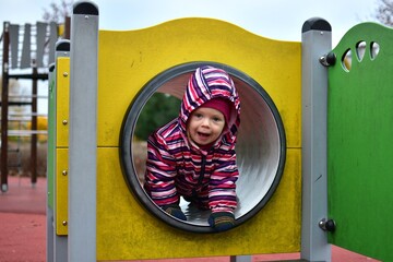 Smiling toddler in a colorful striped snowsuit playing inside a playground tunnel on an autumn day. Bright yellow structure and soft light capture a joyful, candid childhood moment.