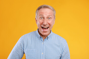 A joyful senior man stands in front of an orange background, laughing heartily while wearing a casual checkered shirt. His expression shows excitement and warmth, enhancing the cheerful atmosphere.