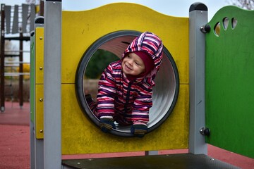Smiling toddler in a colorful striped snowsuit playing inside a playground tunnel on an autumn day. Bright yellow structure and soft light capture a joyful, candid childhood moment.