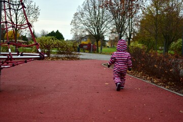 Toddler in a striped snowsuit walking away on a red rubber playground path during autumn. Bare trees, bushes and soft light create a calm, natural outdoor childhood scene.