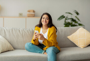 Middle eastern woman sits comfortably on the sofa in her home, smiling at the camera while engaging in a conversation on her smartphone. She appears happy and relaxed in her casual outfit.