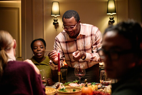 Group of diverse young adults and middle aged Black man celebrating Thanksgiving dinner among friends, smiling and serving food at table with traditional holiday dishes visible