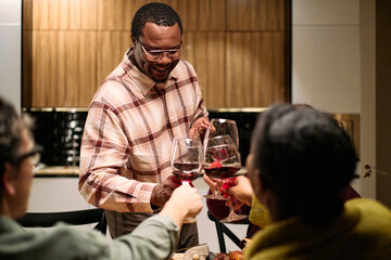 Black middle aged man smiling while serving wine and clinking glasses with diverse group of friends during thanksgiving dinner, visible hands and faces of young and middle aged people participating