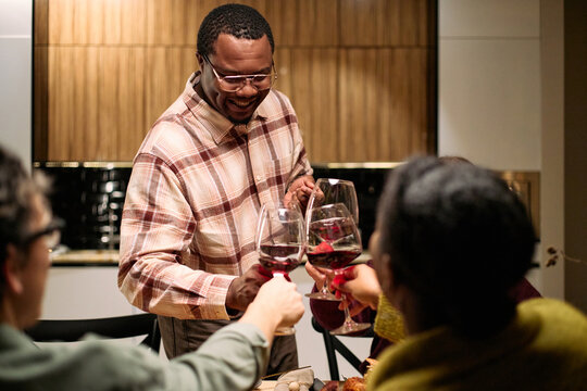 Black middle aged man smiling while serving wine and clinking glasses with diverse group of friends during thanksgiving dinner, visible hands and faces of young and middle aged people participating