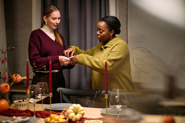 Young adult Caucasian woman and young adult Black woman preparing thanksgiving dinner together, arranging table setting with plates, candles, and festive food, engaging in conversation
