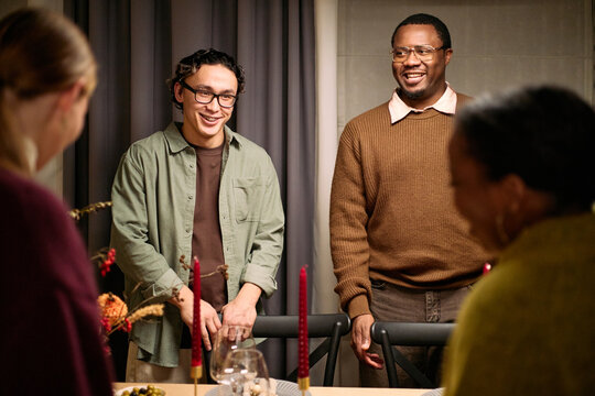 Young adult Asian man and young adult Black man standing and smiling among friends during Thanksgiving dinner, interacting with two women whose faces are partially visible in foreground - Powered by Adobe