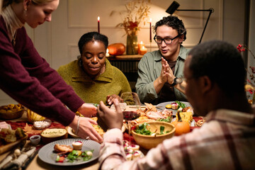Group of diverse young adults and Black woman sitting around table sharing Thanksgiving dinner, eating and talking, hands reaching for food, festive meal setting, autumn decorations visible